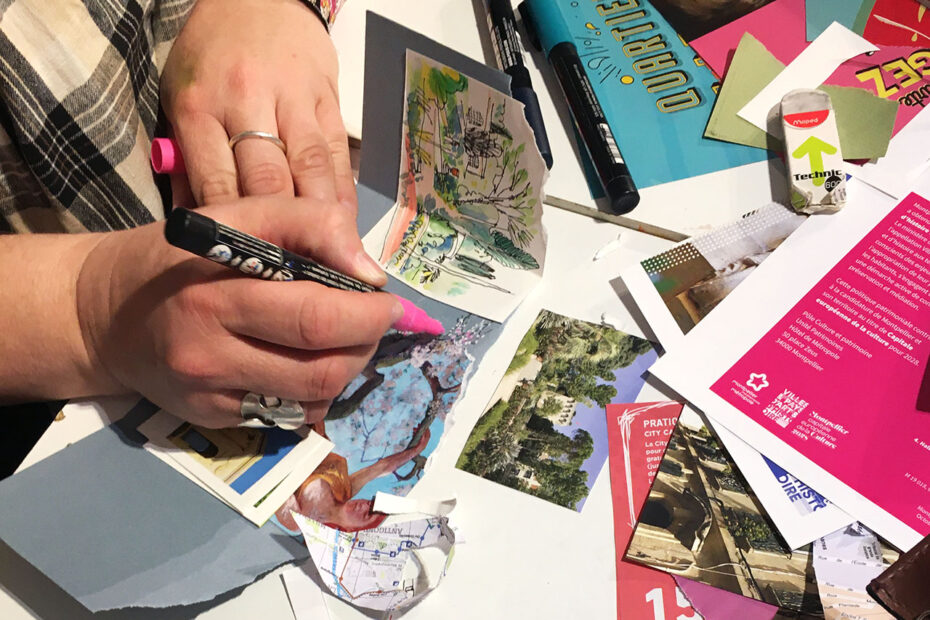Une main dessine au posca sur une page de carnet de voyage à Montpellier, des ciseaux, photos et dépliants touristiques sont étalés sur la table
